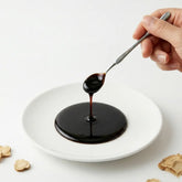 A silver spoon drizzling thick, dark Sahm Canadian Red Ginseng Concentrate onto a white plate with dried ginseng slices on the side.