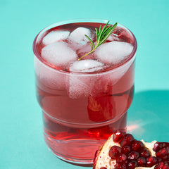 A glass of Danongwon Daily Kombucha Pomegranate with ice and rosemary, placed next to fresh pomegranate pieces on a turquoise background