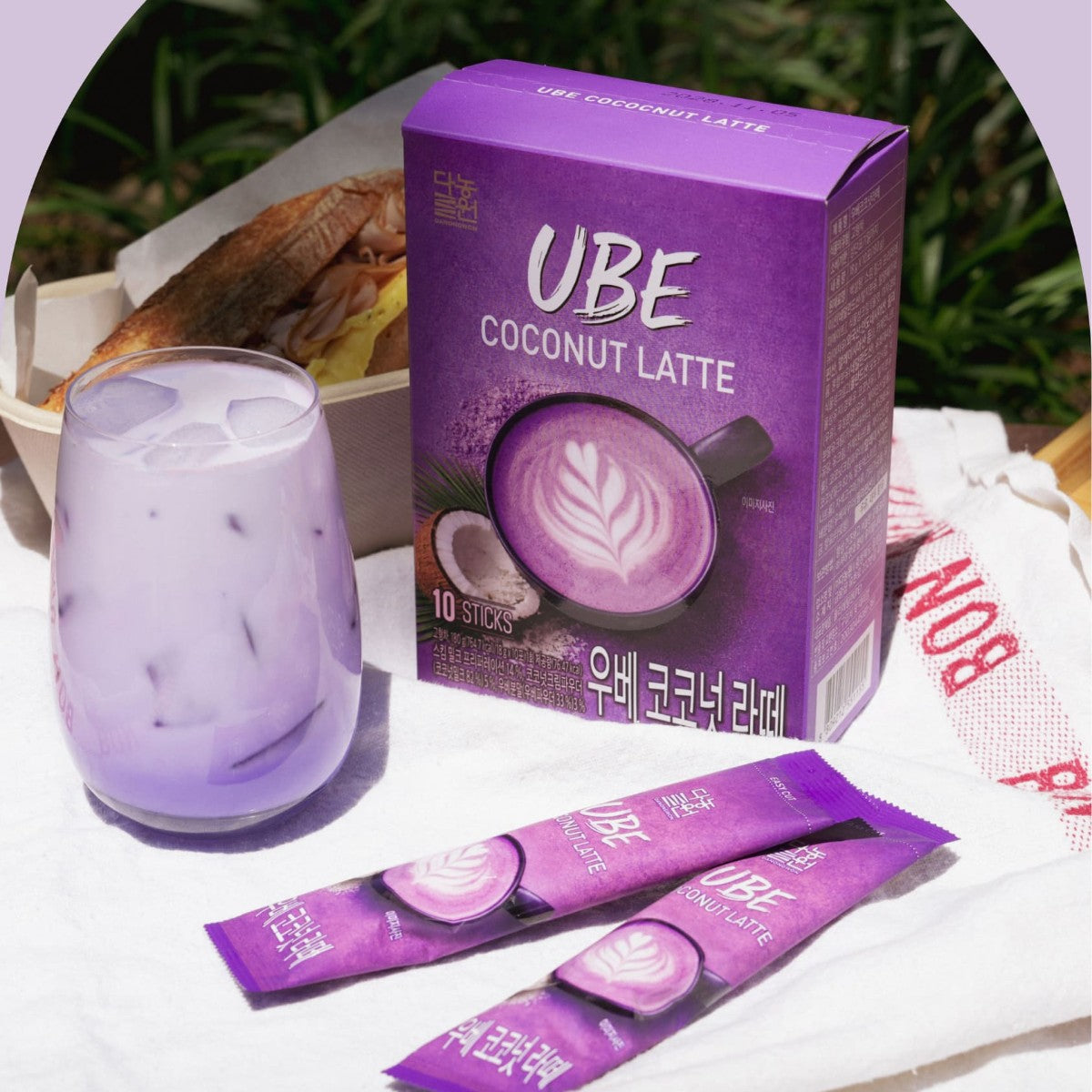 Iced Korean ube coconut latte in a glass with the Danongwon box and two sticks on a picnic cloth