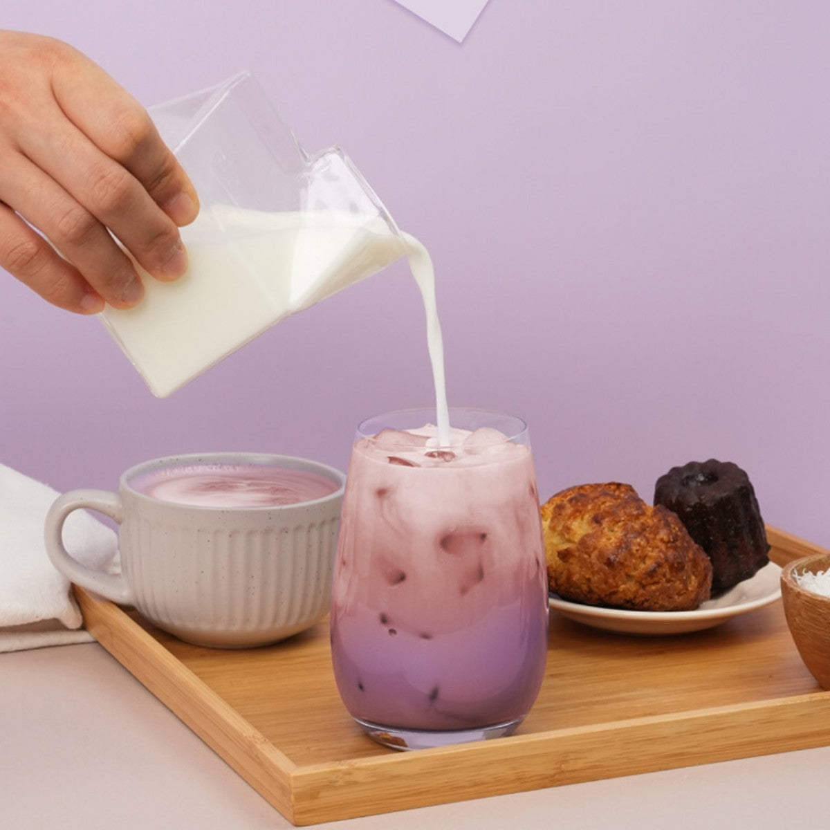 Pouring milk into an iced Korean ube grain latte on a wooden tray with pastries